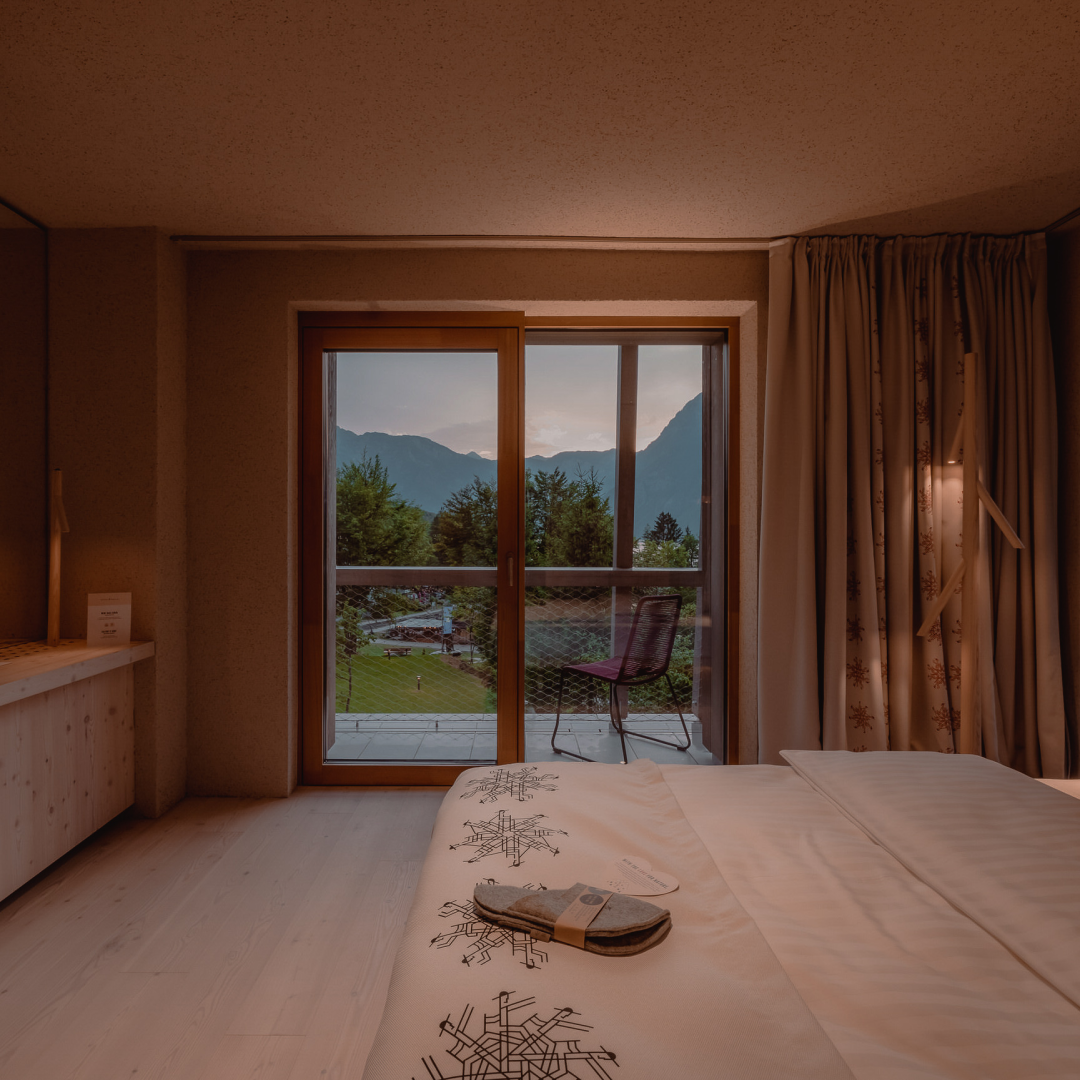 hotel bohinj Udobna hotelska soba s posteljo v ospredju, copati na postelji in steklenimi vrati, ki se odpirajo na balkon s stolom s pogledom na gore in zelenje ob sončnem zahodu.
