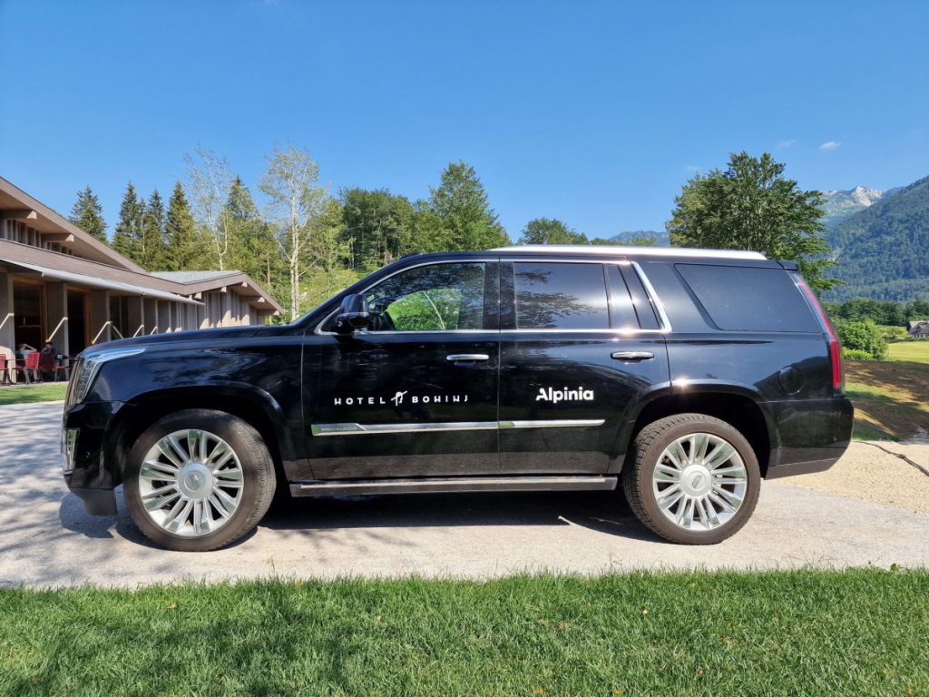 hotel bohinj Črn terenec z logotipoma Hotel Bohinj in Alpinia je parkiran na dovozni cesti ob travnatem območju, v ozadju pa so pod jasnim modrim nebom vidna drevesa, gore in stavba.