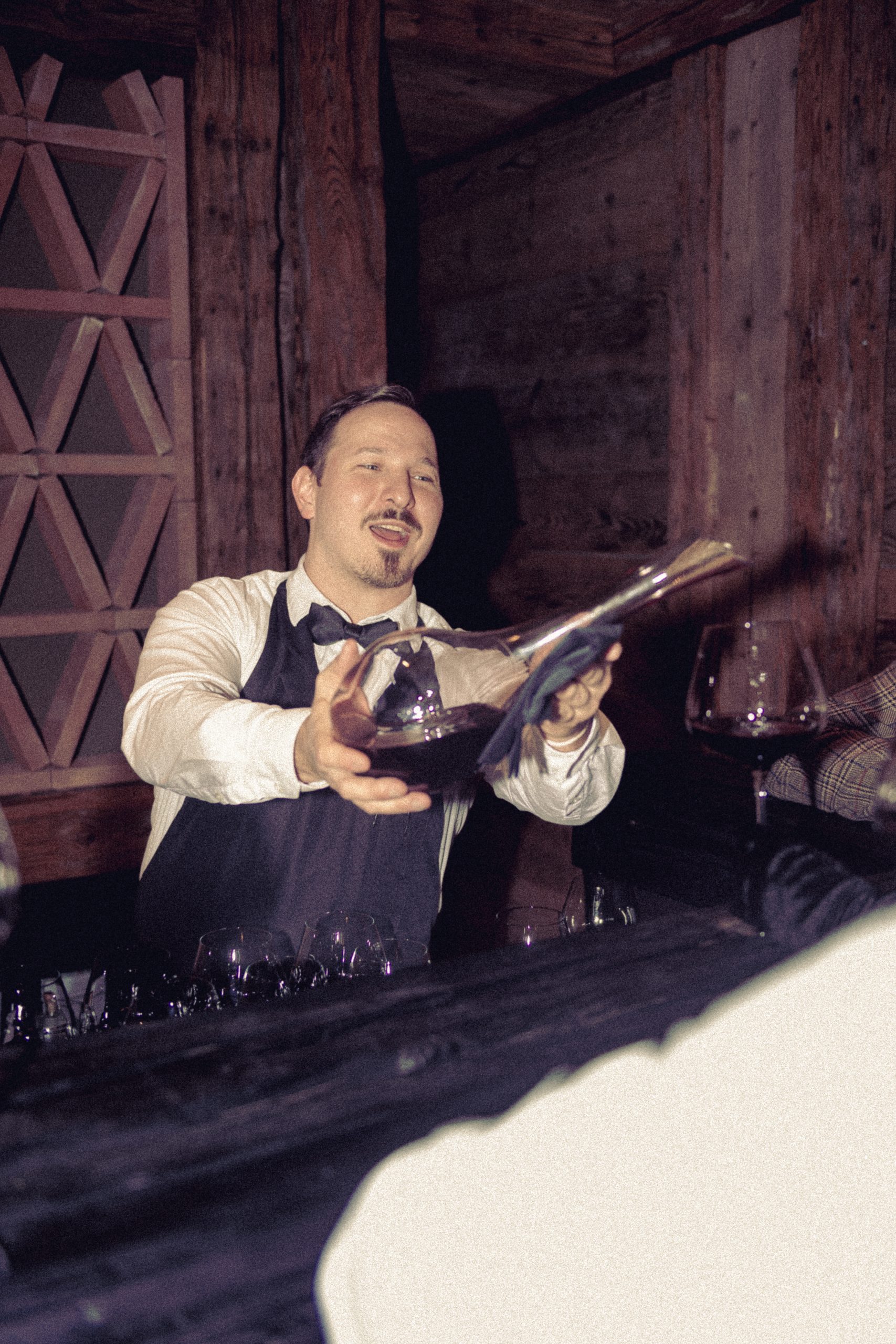 hotel bohinj Barman v metuljčku in predpasniku v rustikalnem lesenem baru z nasmehom preliva vino iz karafe v kozarec.