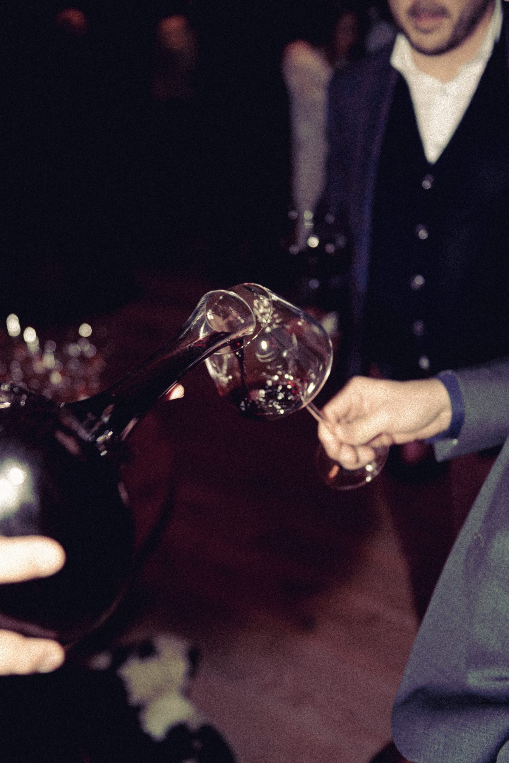 hotel bohinj Oseba iz dekanterja nalije rdeče vino v kozarec, ki ga drži druga oseba, obe oblečeni v formalno obleko na slabo osvetljenem družabnem srečanju.