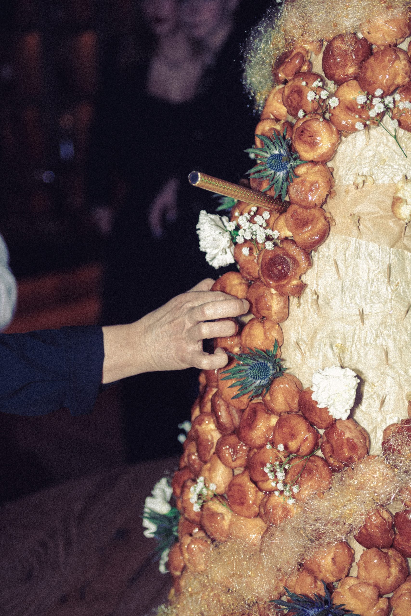hotel bohinj Z roko seže do croquembouche, visoke francoske sladice, ki je narejena iz smetanovih oblivov, zloženih v stožec in okrašenih s sladkorjem, cvetjem in zelenjem. V ozadju so vidne zamegljene figure.