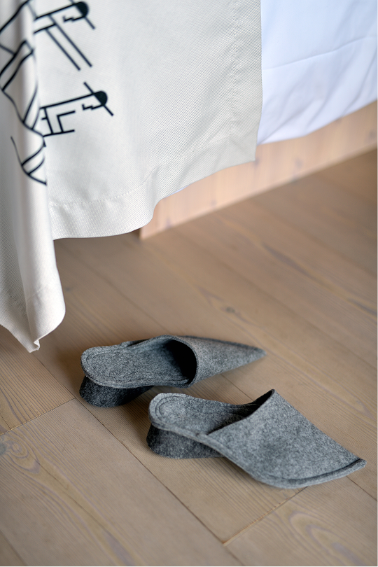 hotel bohinj Na svetlih lesenih tleh ob postelji z belo in vzorčasto pregrinjalo stoji par sivih copat.