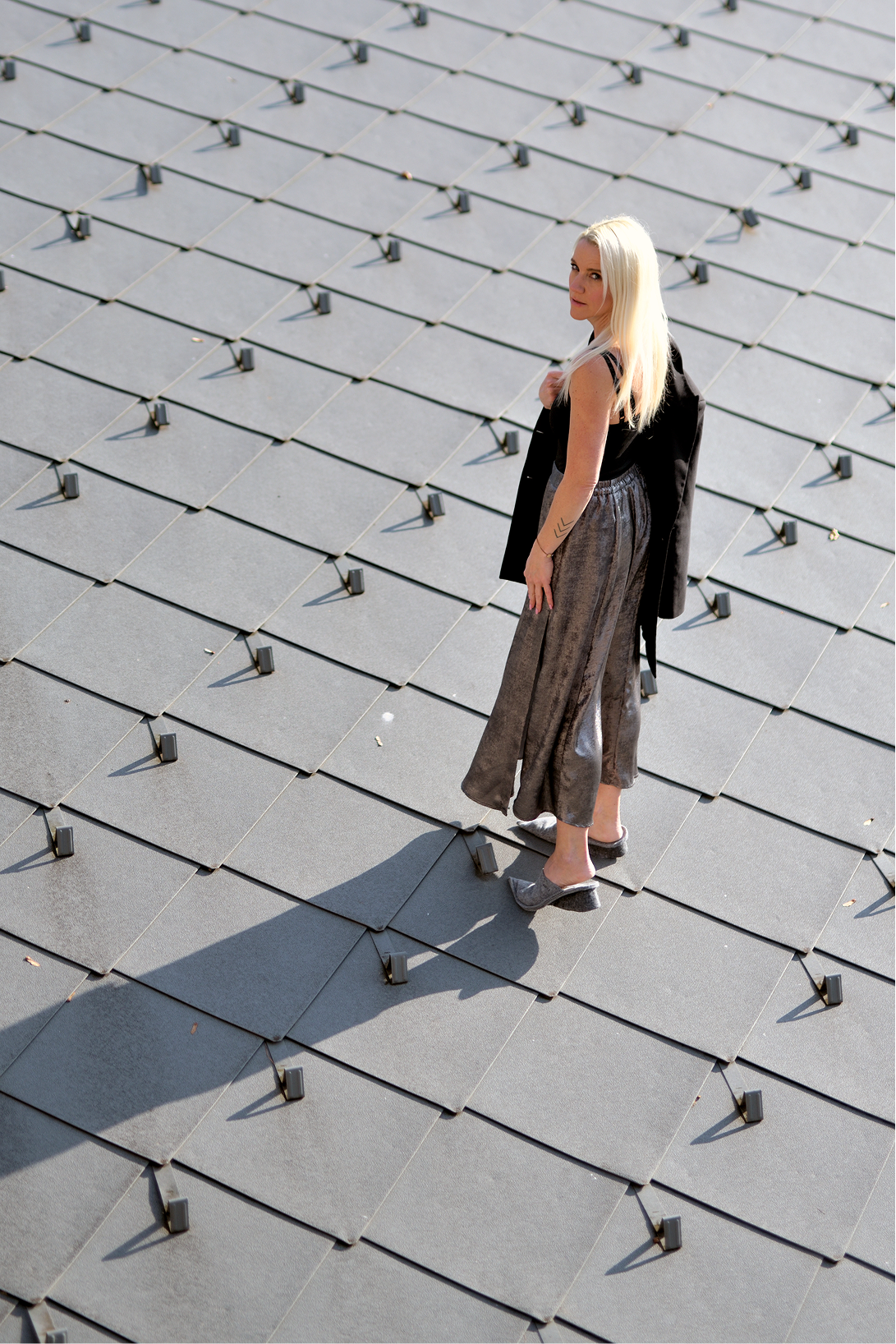 hotel bohinj Ženska z dolgimi svetlimi lasmi stoji na geometrijski površini s ploščicami, oblečena v sivo obleko in s črnim suknjičem čez ramo, ki v sončni svetlobi meče senco.