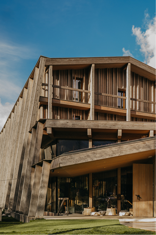 hotel bohinj Sodobna lesena stavba z oglato arhitekturo, velikimi steklenimi okni in odprtimi balkoni na modrem nebu z razpršenimi oblaki in zeleno trato v ospredju.