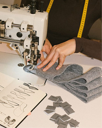 hotel bohinj Oseba, ki s šivalnim strojem šiva kose sive tkanine, z rumenim merilnim trakom okoli vratu. V bližini so nalepke za tkanine in odprt skicirni zvezek z vzorci za šivanje.
